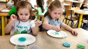 Praca przy stolikach. Dziewczynki malują okrągłe waciki niebieskim i zielonym barwnikiem przy pomocy pipety.