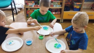 Chłopiec wkrapla pipetą niebieski barwnik na talerz z zielonymi cukierkami.